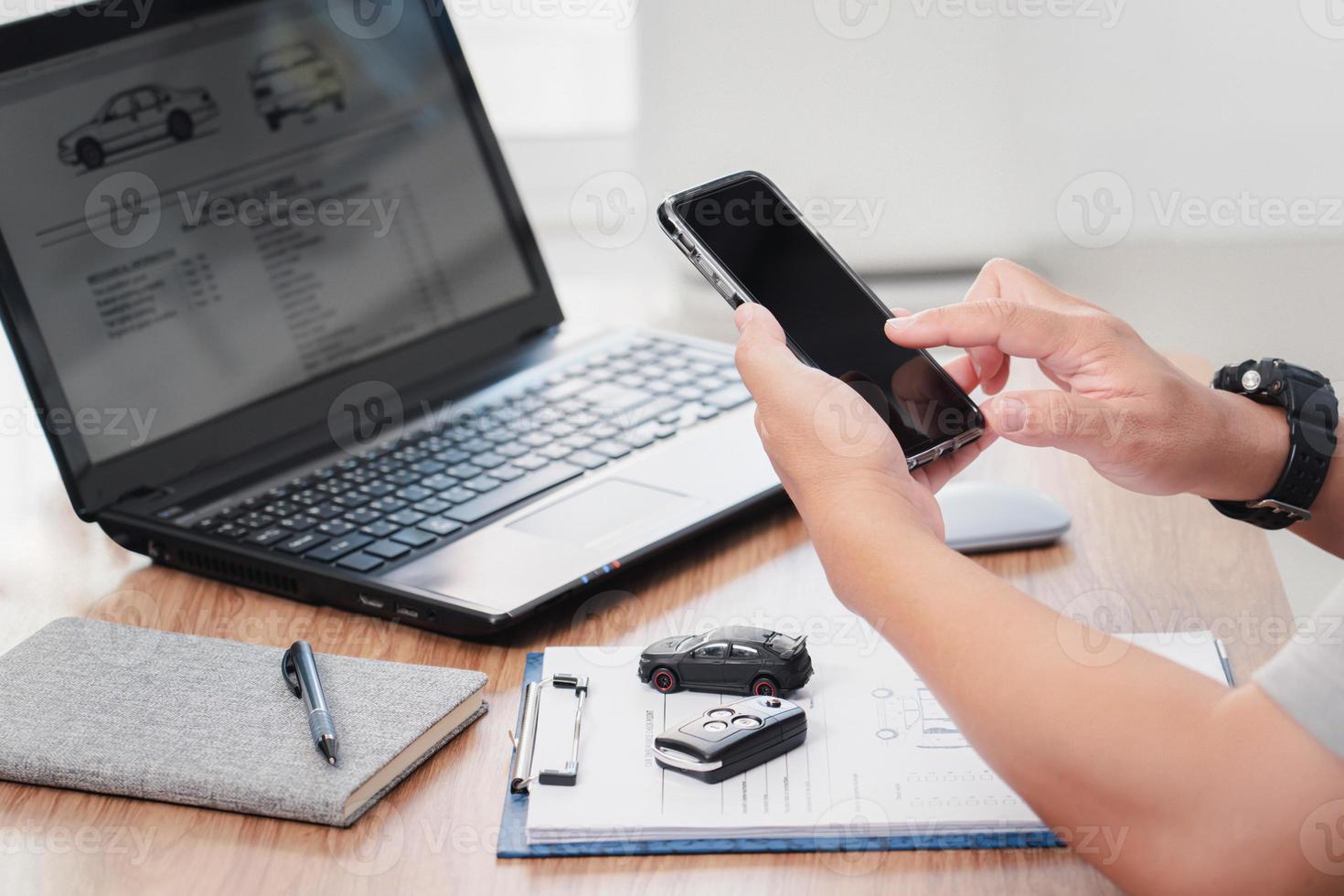 Car insurance online concept , A man hold smartphone and with checklist car key toy car on table and use laptop check online case of accident photo