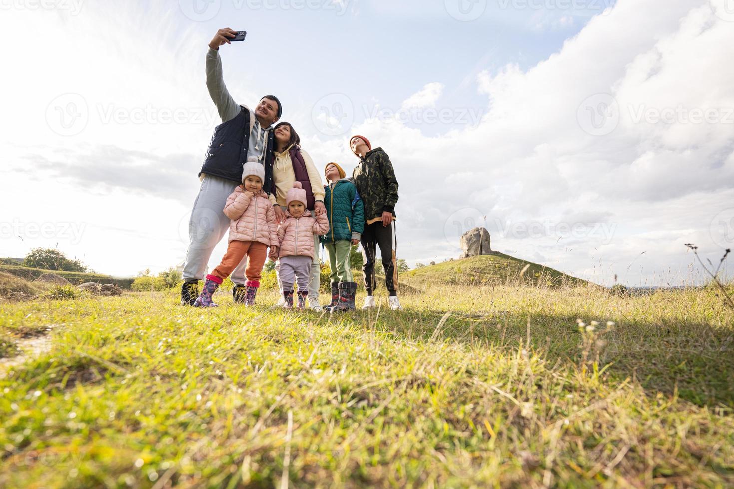 Family exploring nature and making selfie on phone. Children with parents hiking against big stone in hill. Pidkamin, Ukraine. photo
