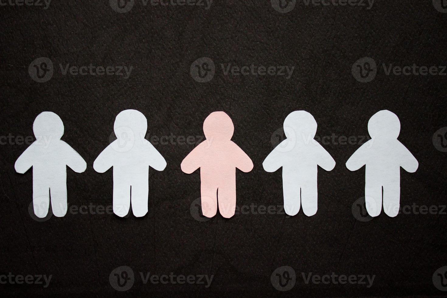 Silhouettes of five men, arranged in a row, cut from paper. Between the white men there is one pink man. On black background photo