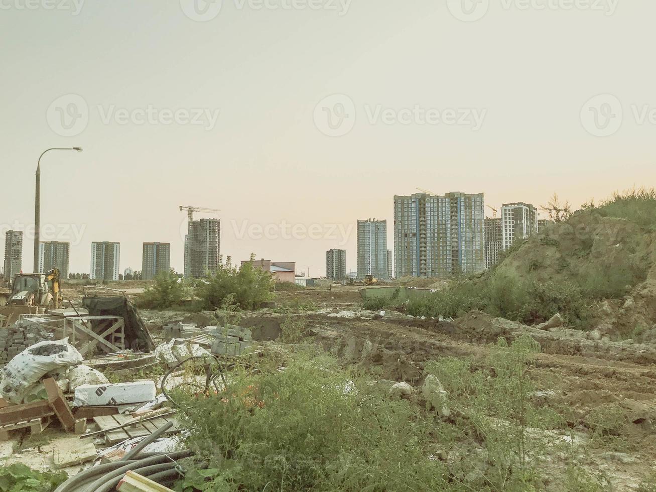 industrial site. construction of a large residential complex. nearby are green bushes where construction materials and debris lie. built concrete houses in the distance photo