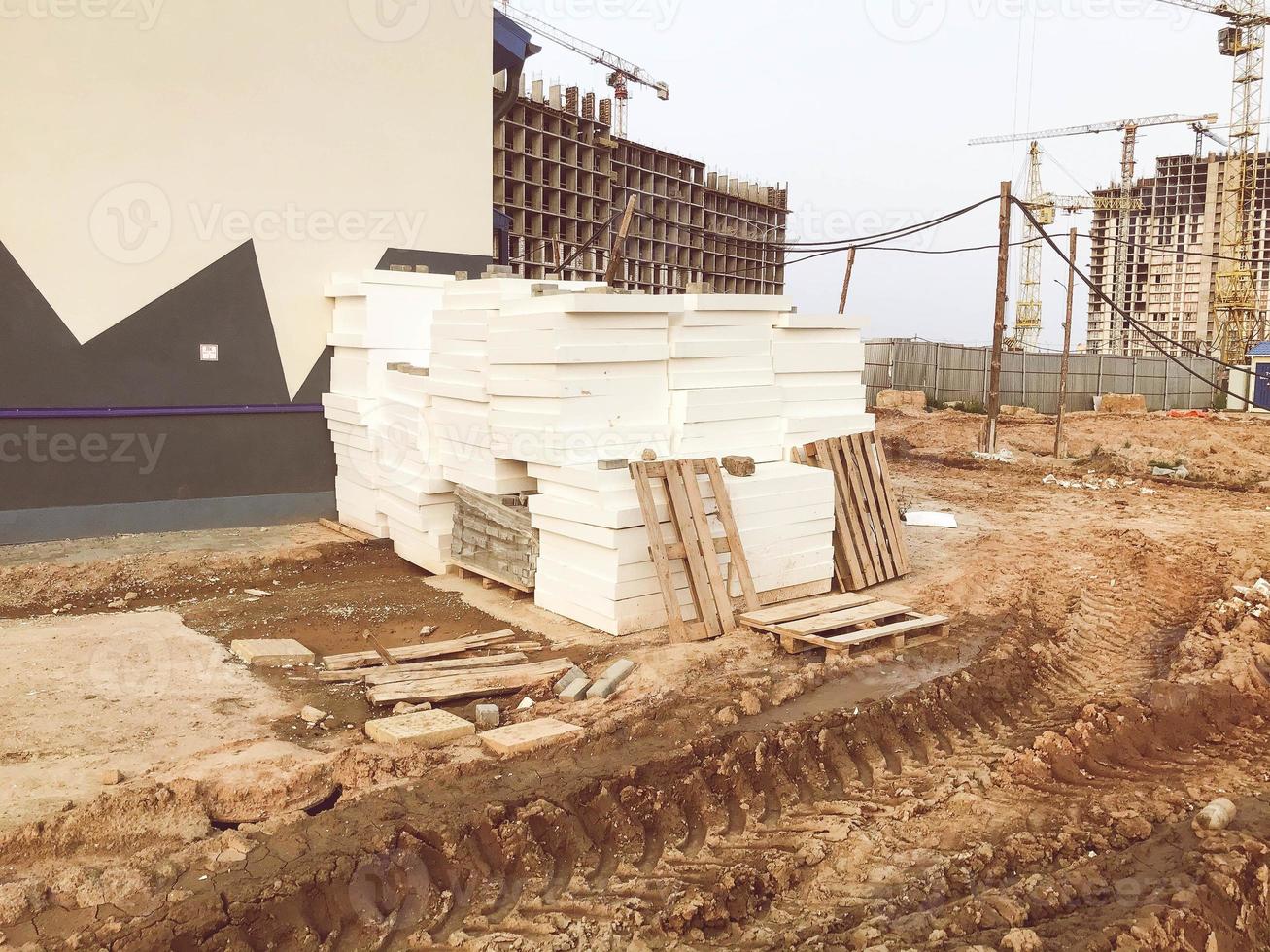 building materials are piled up near the building. white blocks are packed in foil. next to wooden pallets, unfinished houses. traces and furrows of large vehicles on the sand photo