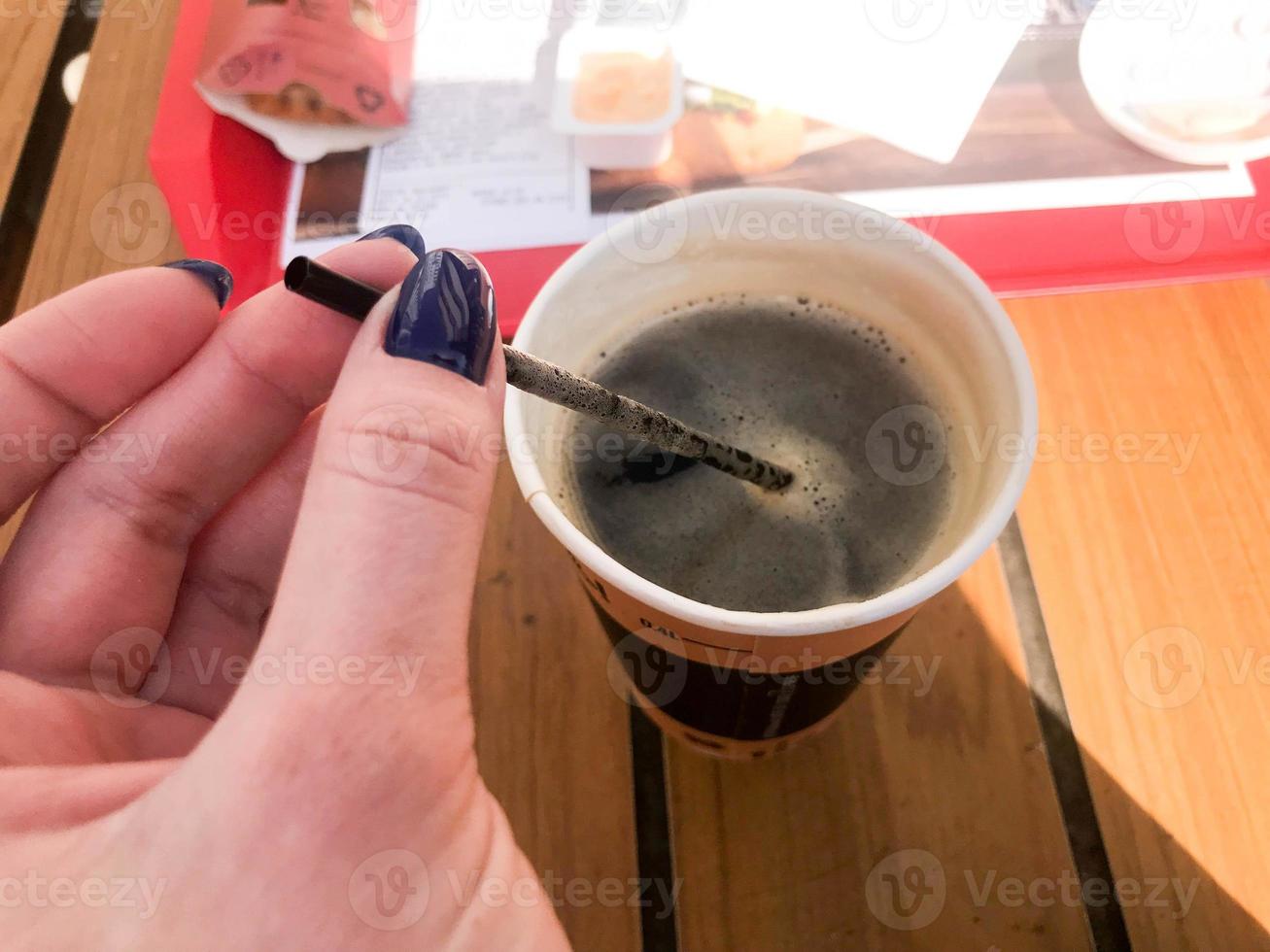 una mujer sostiene con una hermosa manicura en los dedos un tubo en una taza de café natural rápido, negro y caliente de comida rápida en un vaso de papel foto