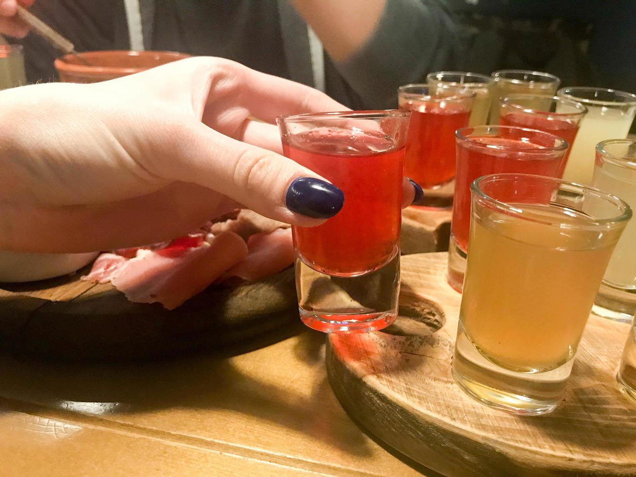 Woman, girl holds in her hand with a manicure on her fingers a delicious red glass, a shot with strong alcohol, vodka, brandy, brandy on wooden stands on a table in a cafe, bar, restaurant photo