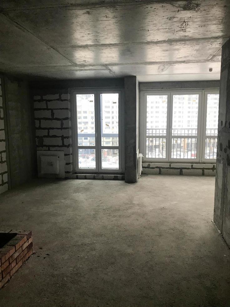 The apartment in the new building with a free layout without repair and decoration with bare walls and a large panoramic French window on the whole wall. Concept repair, housewarming, building photo
