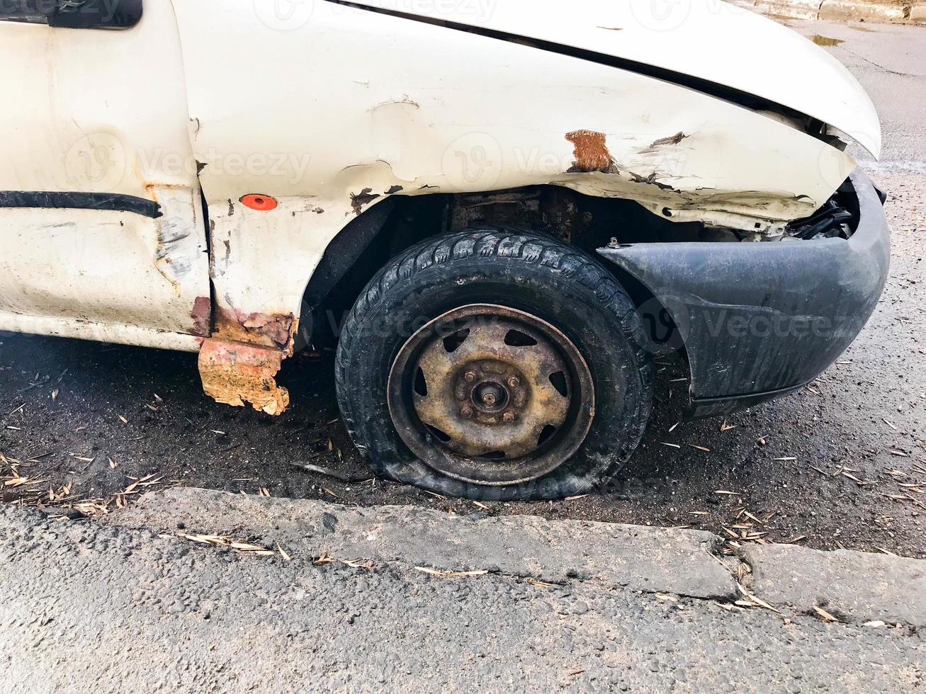 Old white rusty broken car of the carcass with lowered punctured wheels with scratches corrosion and a torn-off bumper with dents and scratches thrown on the road. Auto junk photo