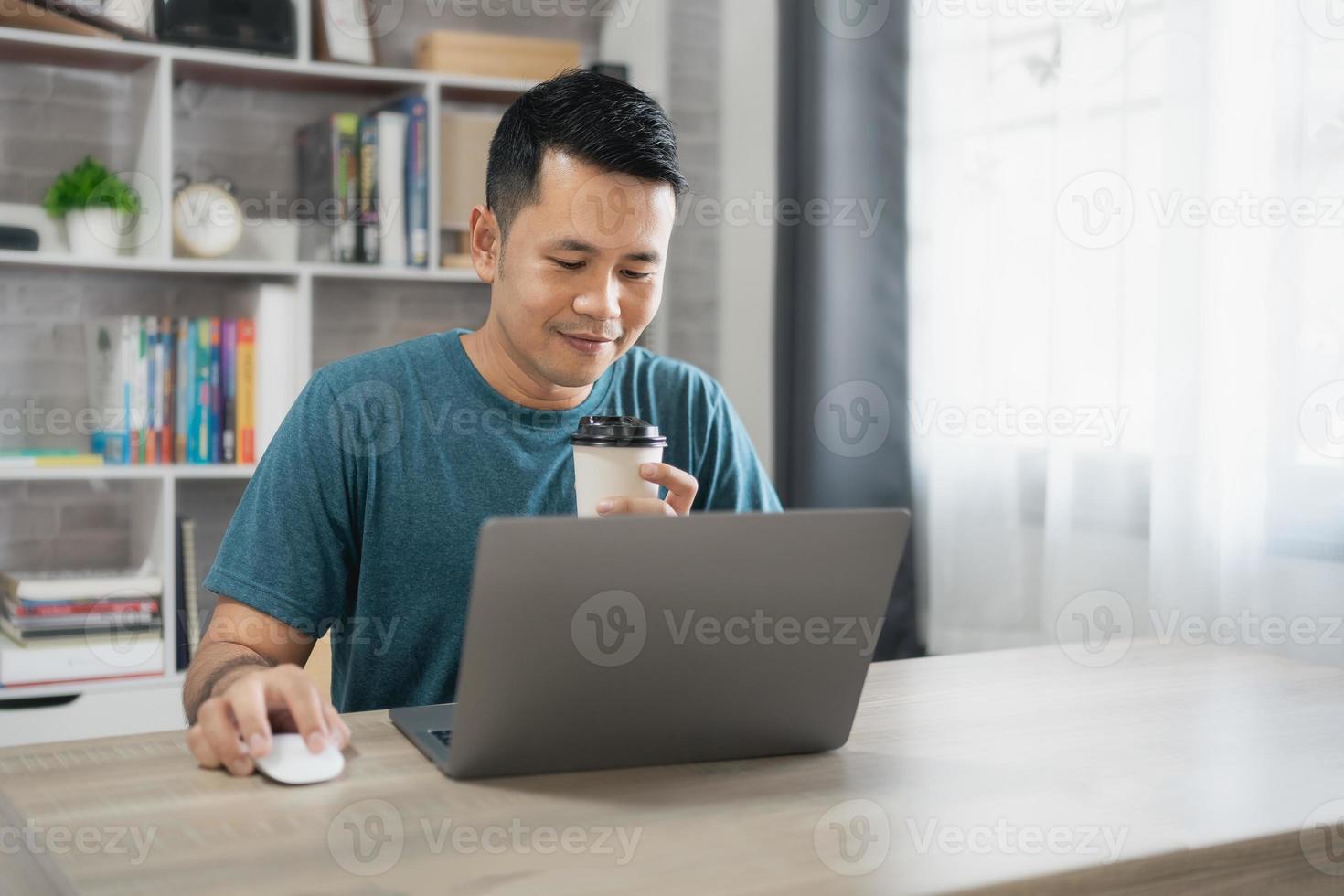 Asian freelance man smiling holding cup of hot coffee and working on laptop computer on wooden table at home. Entrepreneur man working for his business at home. Business work at home concept. photo