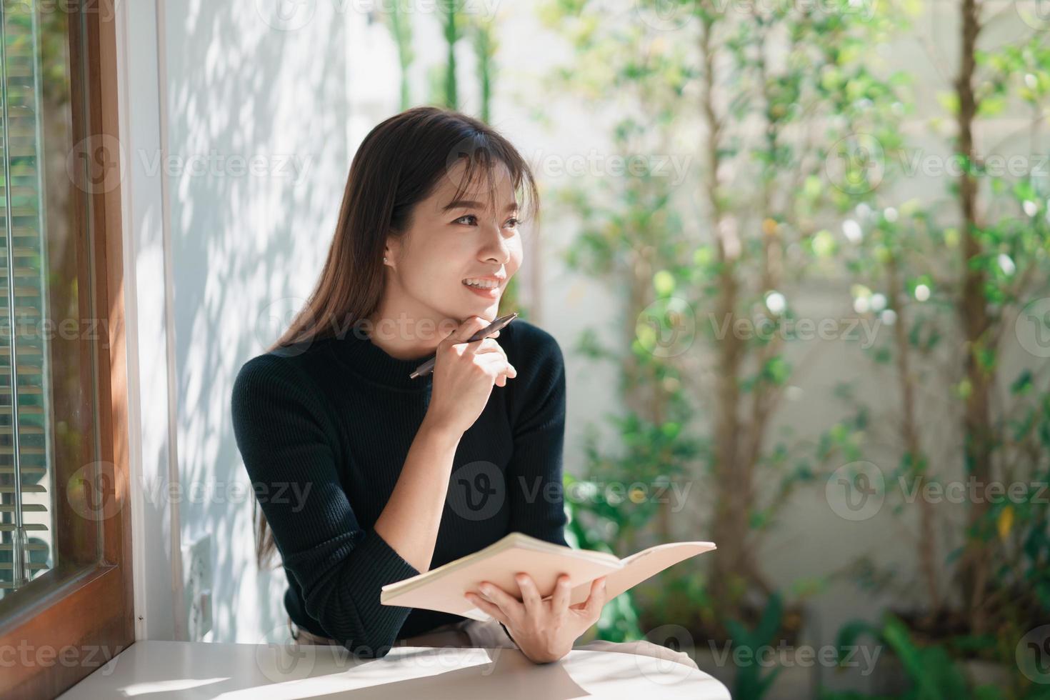 Asian working woman wearing black shirt and writing journal on small notebook on the table at indoor cafe. Woman notes and drinking coffee at cafe. Working from anywhere concept. photo