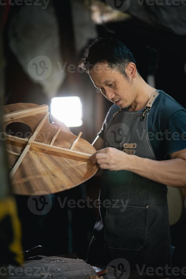 fabricantes de guitarras hombre asiático haciendo guitarras acústicas en laboratorio. fabricante de guitarras asiático construye guitarras de alta calidad para músicos tienda de guitarras hechas a mano. trabajo en madera fina, tradición, artesanía milenaria. foto