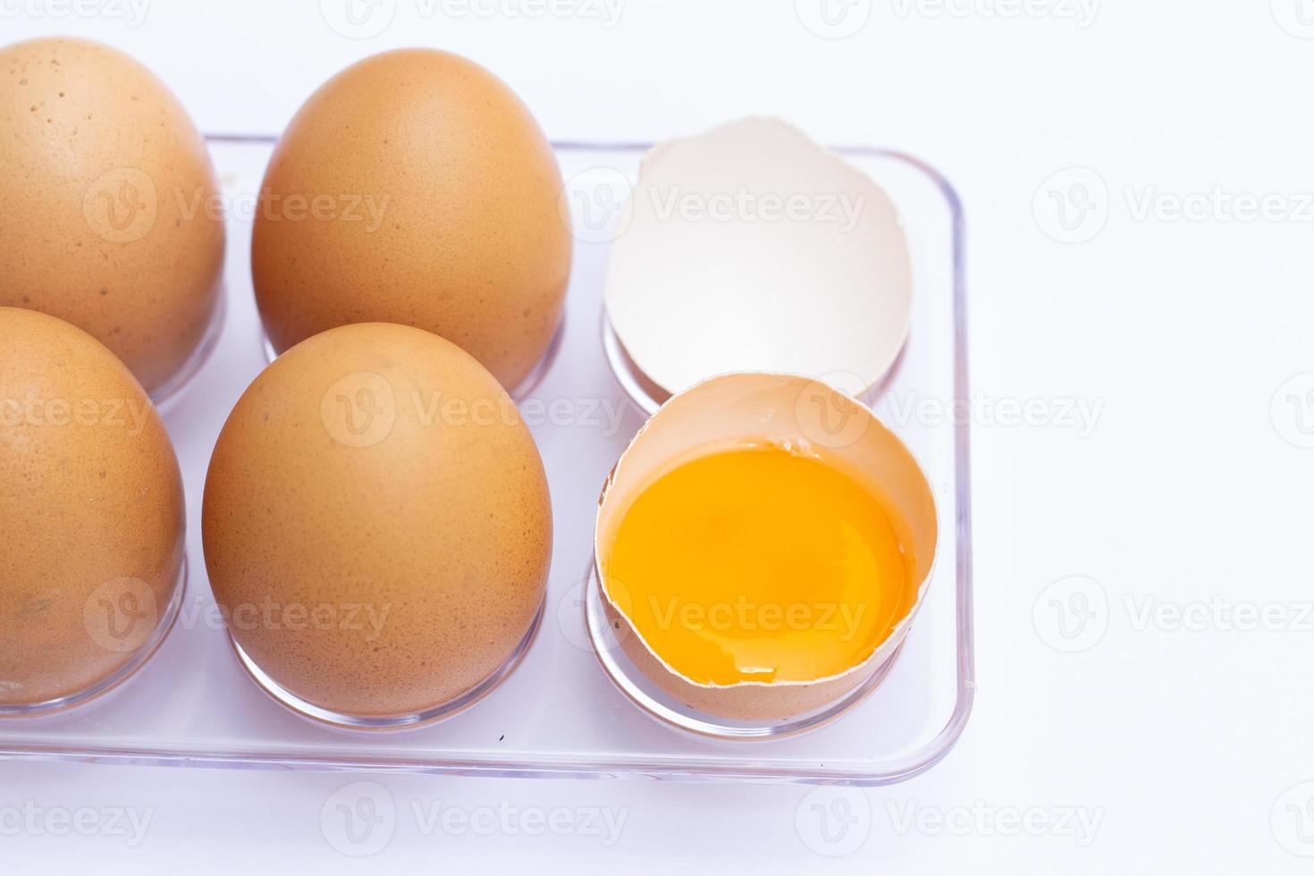 cuatro huevos marrones yacían en una bandeja de huevos con una cáscara de huevo sobre un fondo blanco. había un huevo roto, mostrando la yema adentro. foto