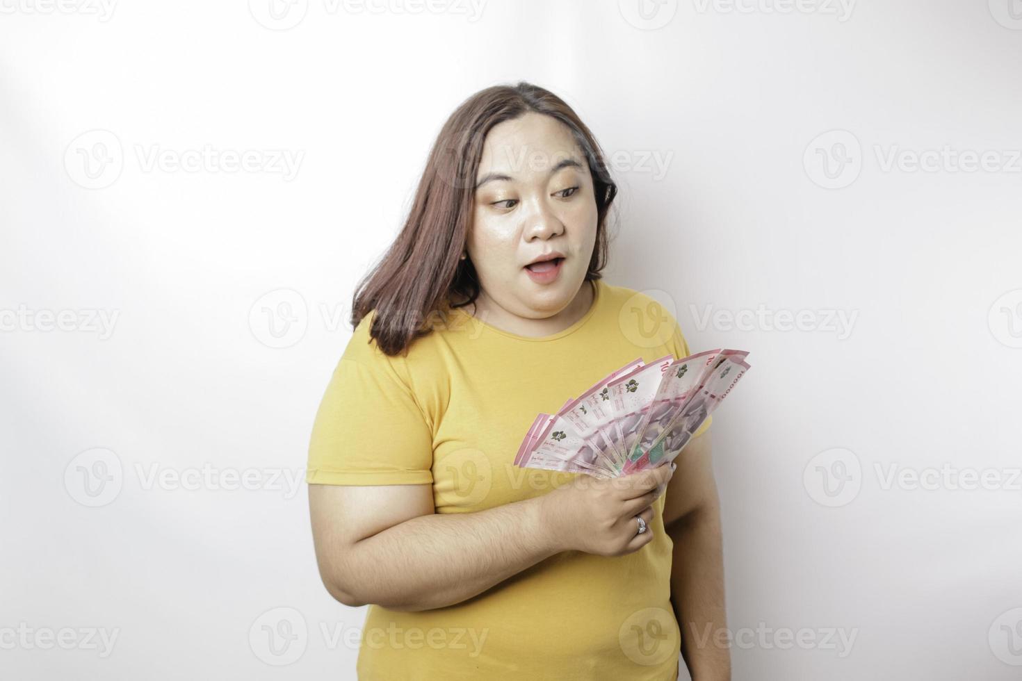 una mujer joven y feliz de gran tamaño lleva una camiseta amarilla y tiene dinero en efectivo en rupias indonesias aislada de fondo blanco foto