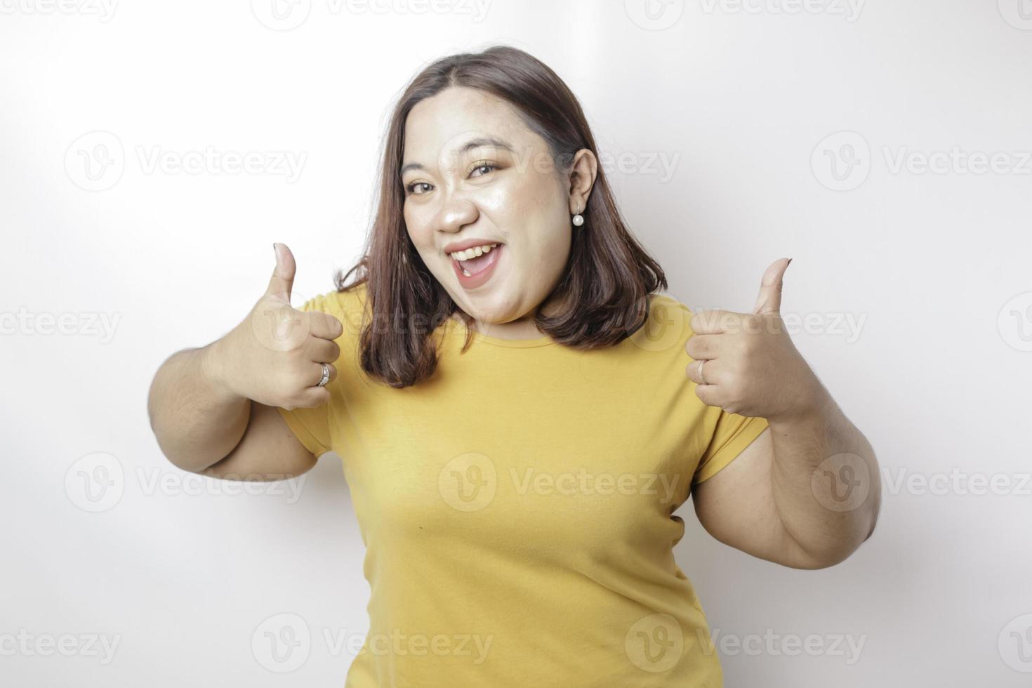 Excited Asian big sized woman wearing yellow shirt gives thumbs up hand gesture of approval, isolated by white background photo