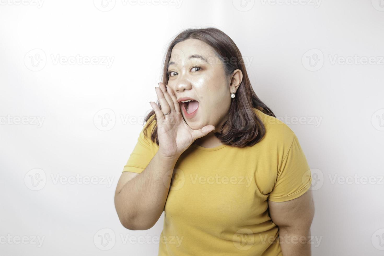 joven y hermosa mujer asiática de gran tamaño con una camisa amarilla gritando y gritando fuerte con una mano en la boca. concepto de comunicación foto