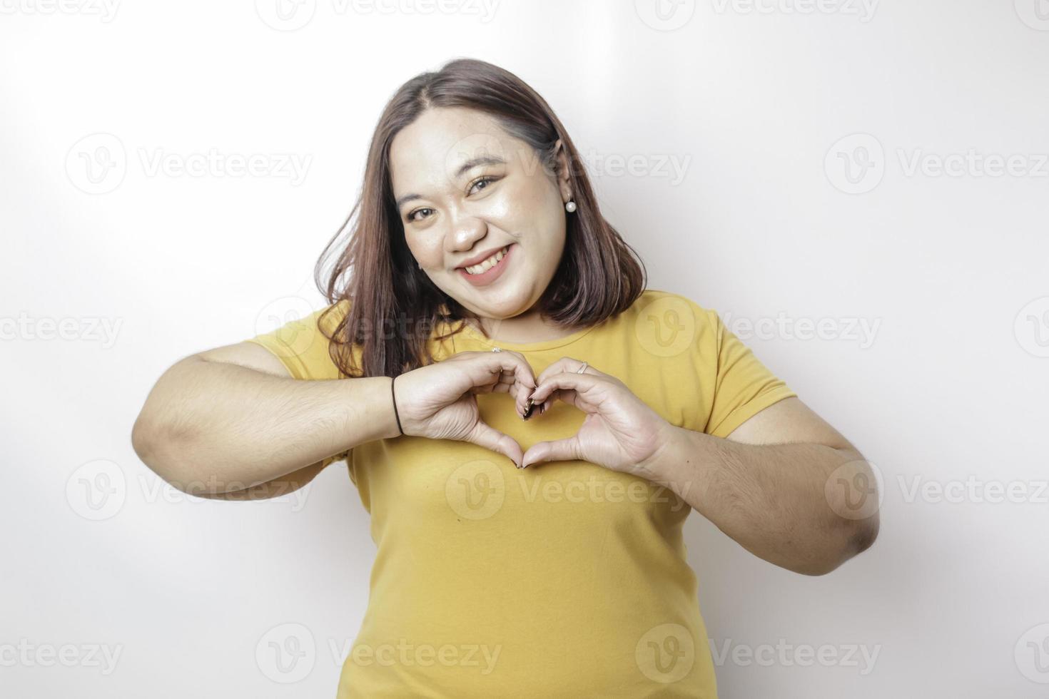 An attractive young Asian big size woman wearing a yellow t-shirt feels happy and a romantic shapes heart gesture expresses tender feelings photo