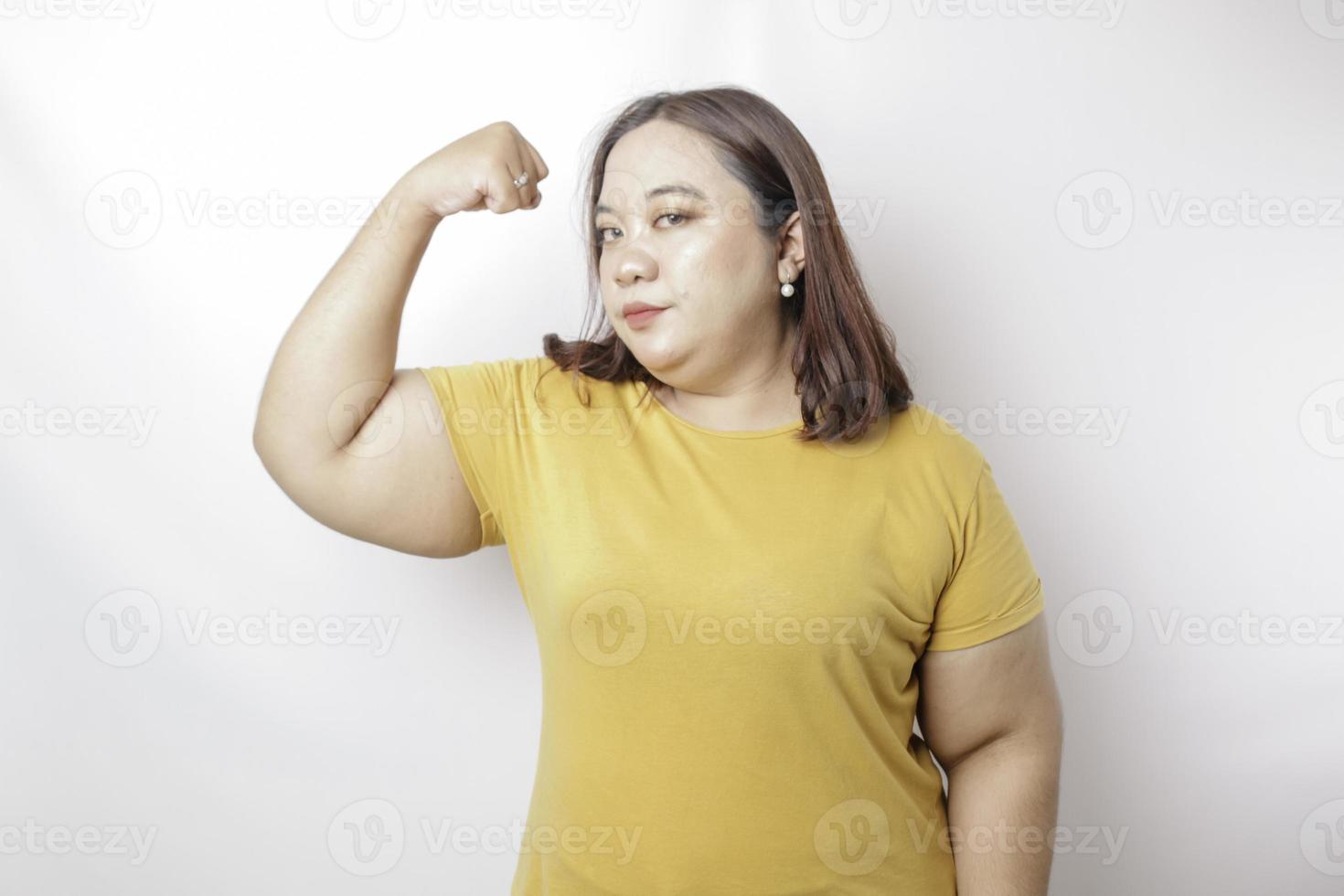 Excited Asian big sized woman wearing a yellow t-shirt showing strong gesture by lifting her arms and muscles smiling proudly photo