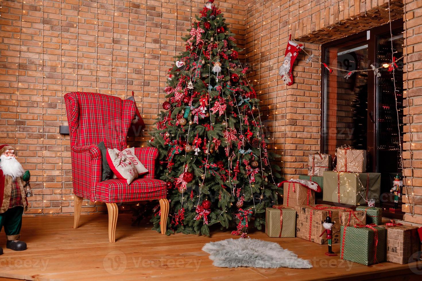 Room decorated to christmas celebration. christmas tree with red balls, bows, gift boxes near the window, cozy chair and santa near brick wall photo