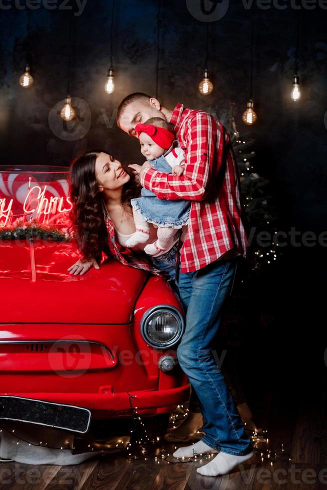 Young beautiful parents holding their little cute daughter in their arms having fun near retro car in studio. Christmas family look. New Year's scenery. photo