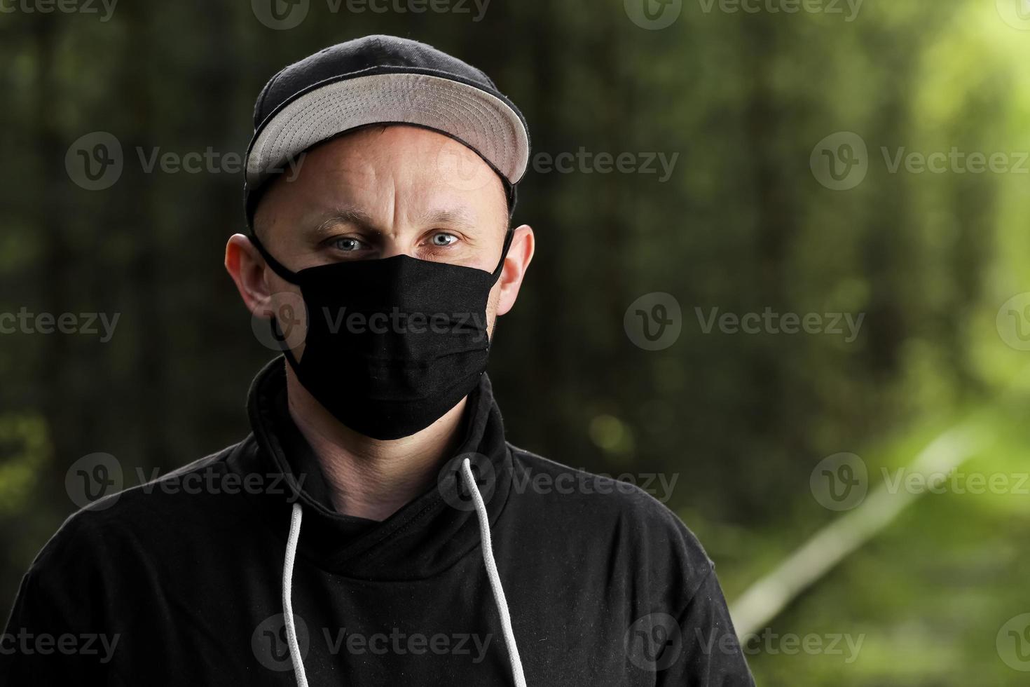 Young man in black protective antiviral mask in the summer park. The guy is resting outdoors on a sunny summer day in a mask made by his own hands. Virus protection. Quarantine measures photo
