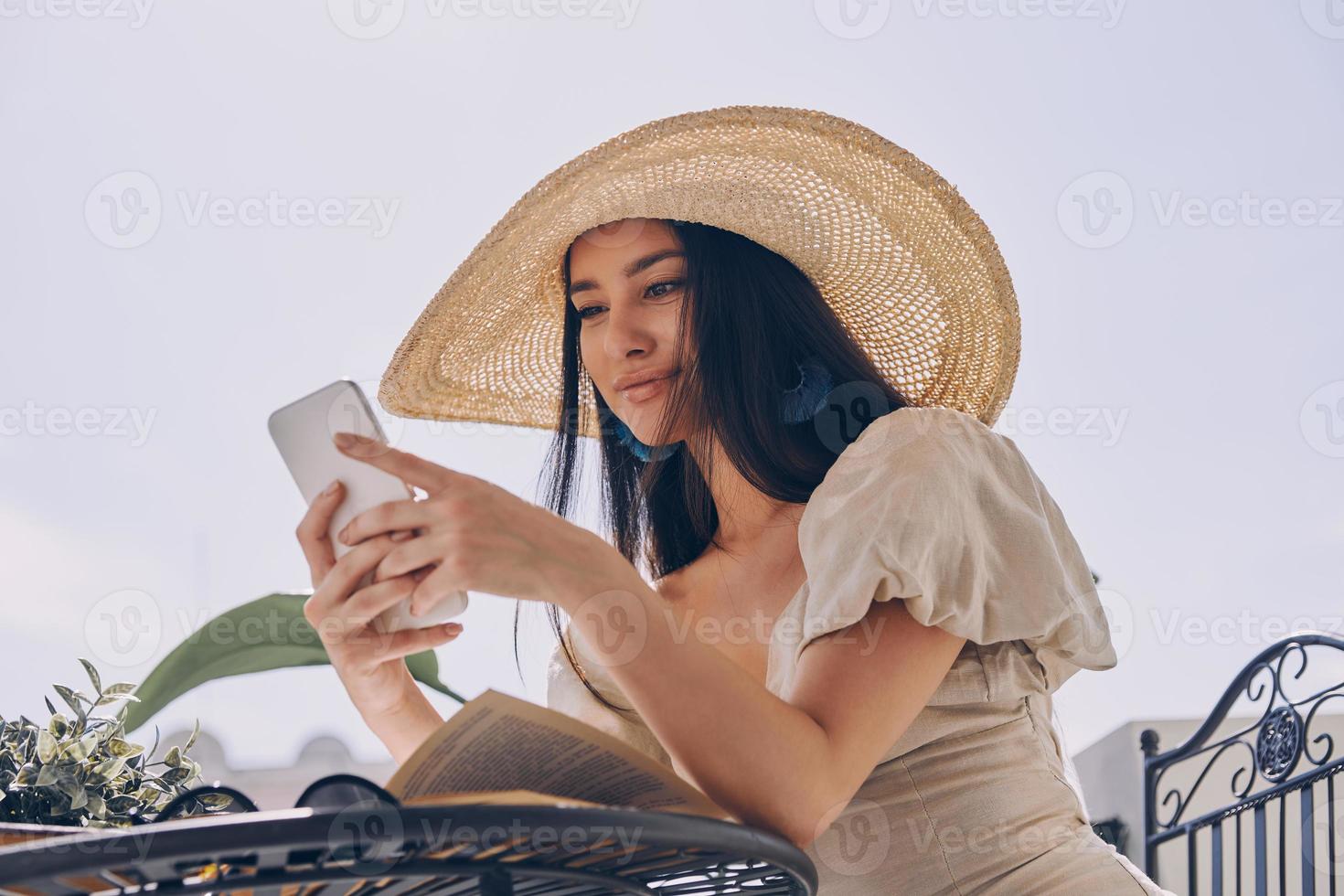 bella joven con sombrero elegante usando un teléfono inteligente mientras se relaja en el balcón foto