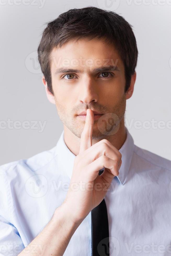 Keep it between us Confident young man in white shirt looking at camera and holding finger on lips while standing against grey background photo