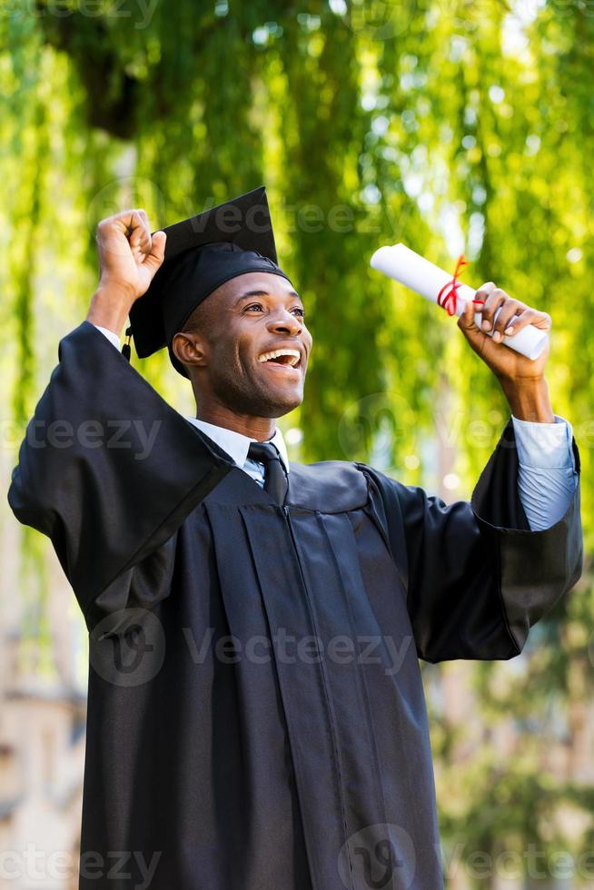 finalmente se graduó feliz joven africano en vestidos de graduación con diploma y levantando los brazos foto