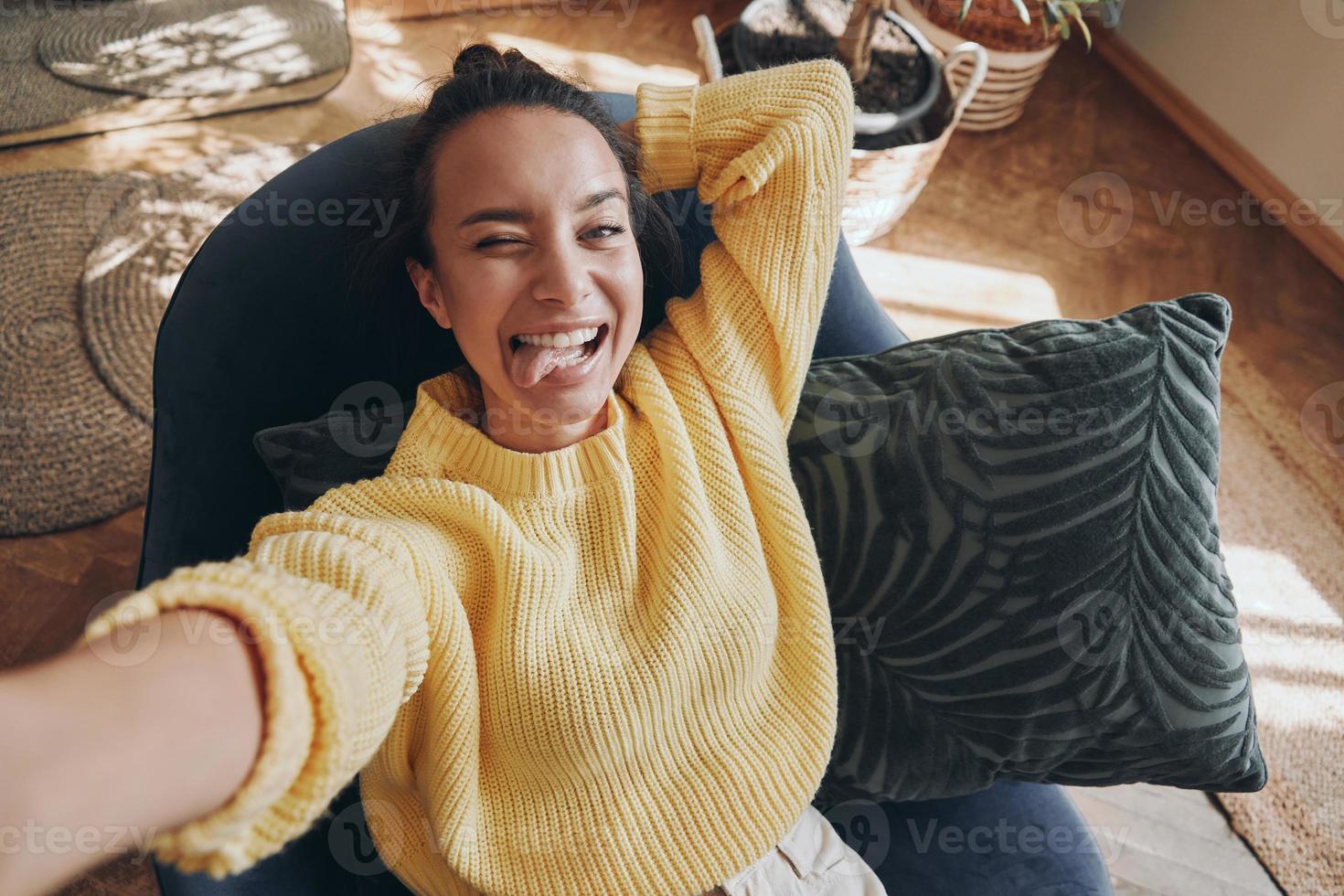 Top view of playful young woman making selfie and grimacing while relaxing in chair at home photo