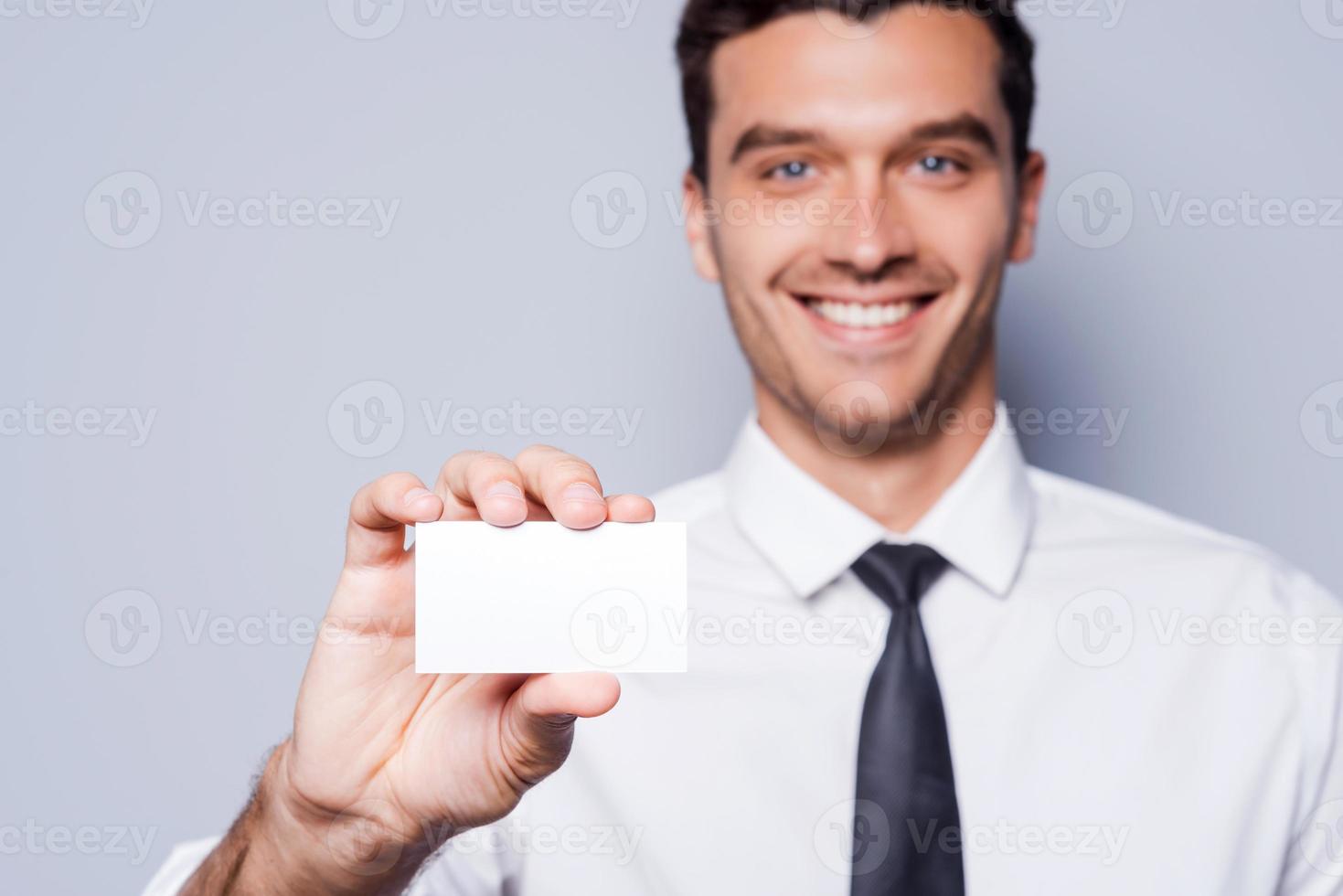 llámame en cualquier momento apuesto joven con camisa y corbata mostrando su tarjeta de visita y sonriendo mientras está de pie contra el fondo gris foto