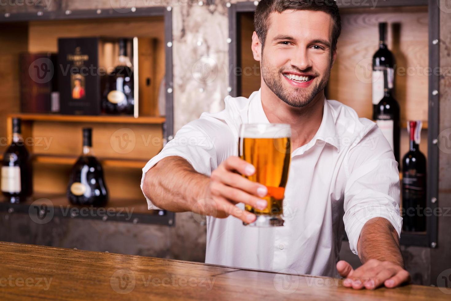 Let me quench your thirst Happy young male bartender in white shirt stretching out glass with beer and smiling while standing at the bar counter photo