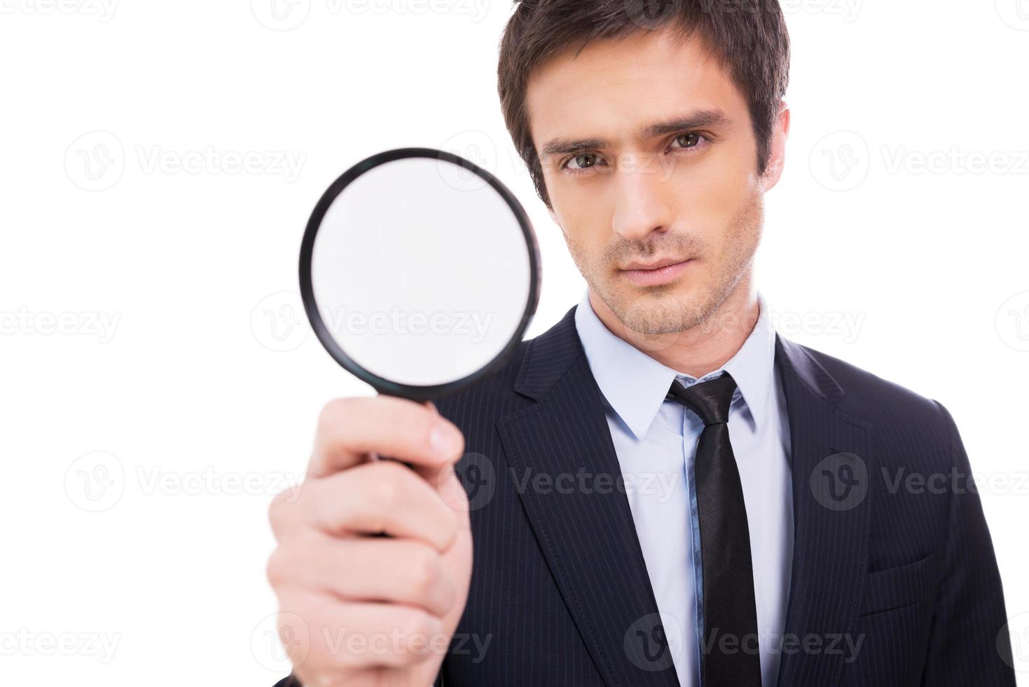 I need details Concentrated young man in formalwear holding magnifying glass and looking at camera while standing isolated on white background photo
