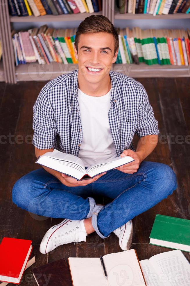 I love studying Top view of happy young man reading book and looking at camera while sitting against bookshelf photo