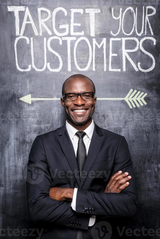 Target your customers Happy young African man in formalwear keeping arms crossed while standing against blackboard with sketch on it photo