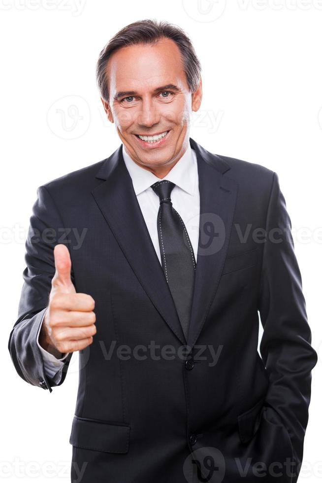 Good job Confident mature man in formalwear looking at camera and smiling while showing his thumb up and standing against white background photo