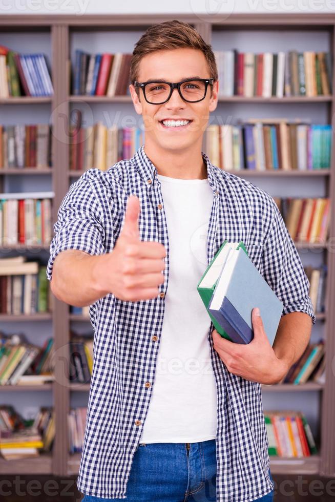 me encanta estudiar a un joven feliz sosteniendo libros y mostrando su pulgar hacia arriba mientras está parado en la biblioteca foto