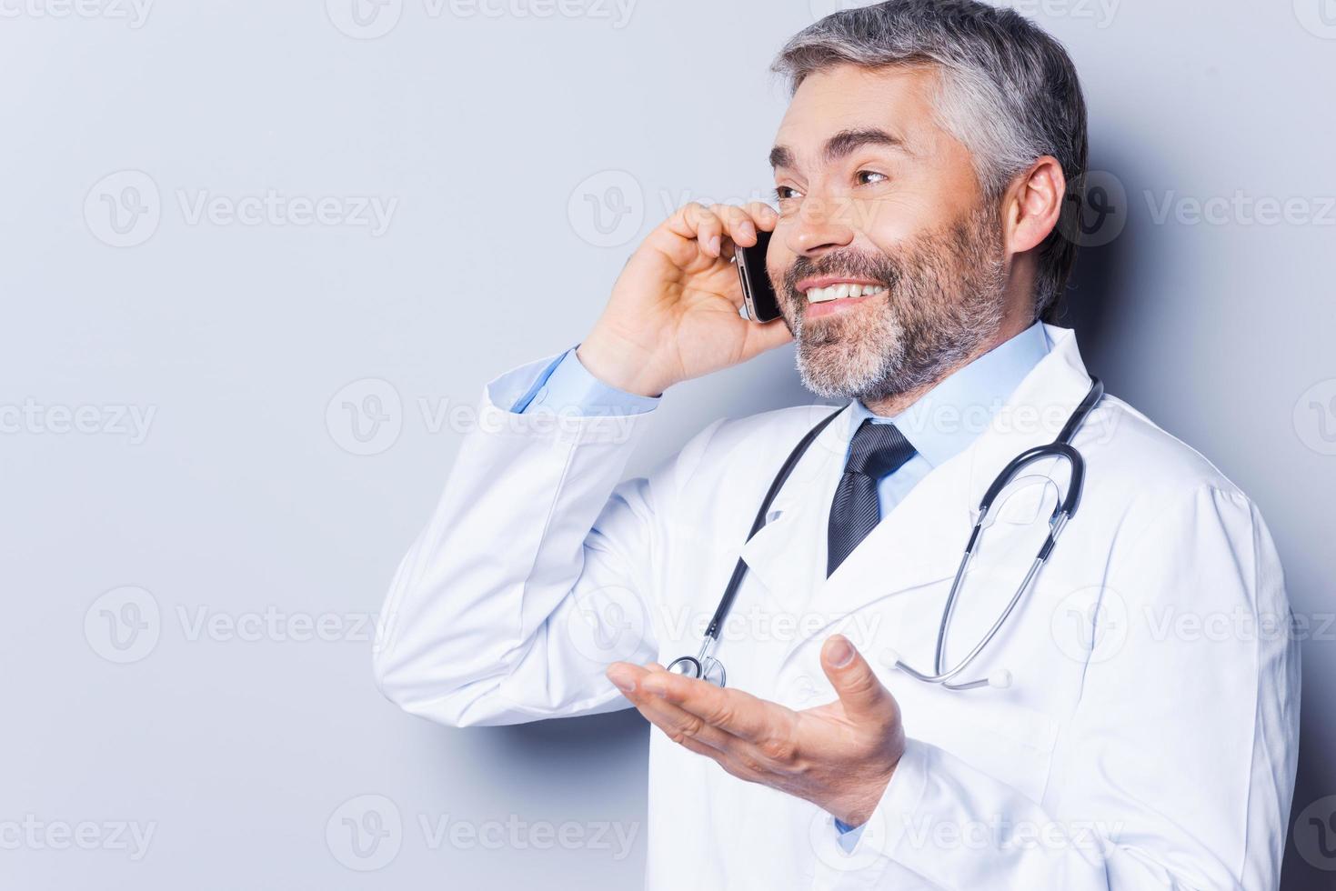 Great news Happy mature grey hair doctor talking on the mobile phone and gesturing while standing against grey background photo