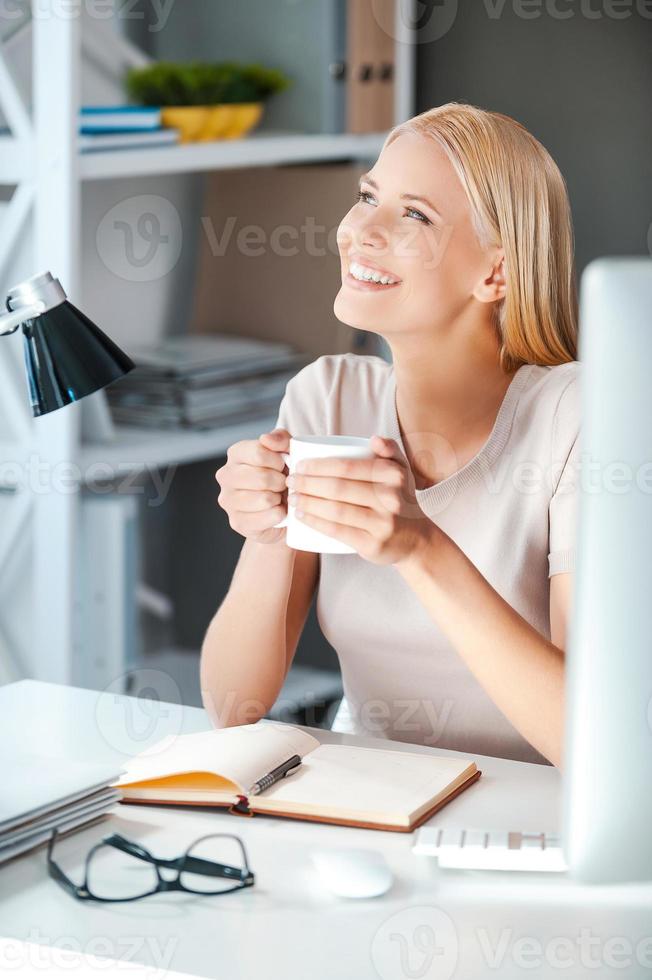 qué hermoso día hermosa mujer joven sosteniendo una taza con bebida caliente y mirando hacia otro lado con una sonrisa mientras se sienta en su lugar de trabajo en la oficina foto