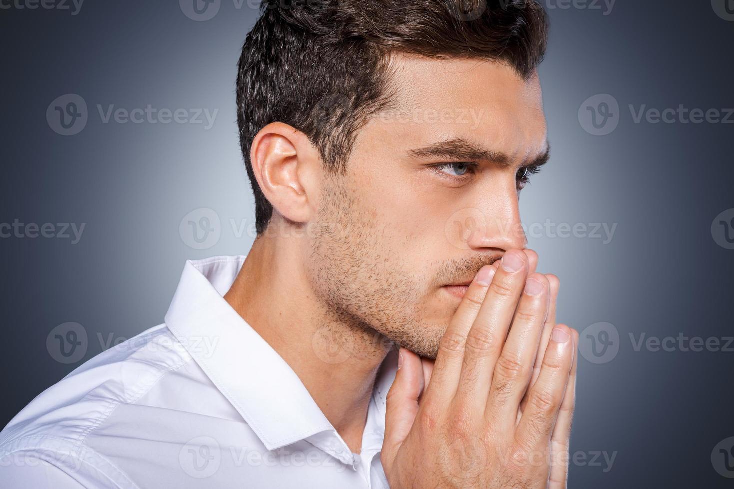 Concentrating at Side view of concentrated young man in white shirt holding hands clasped near face and looking away while standing against grey background photo