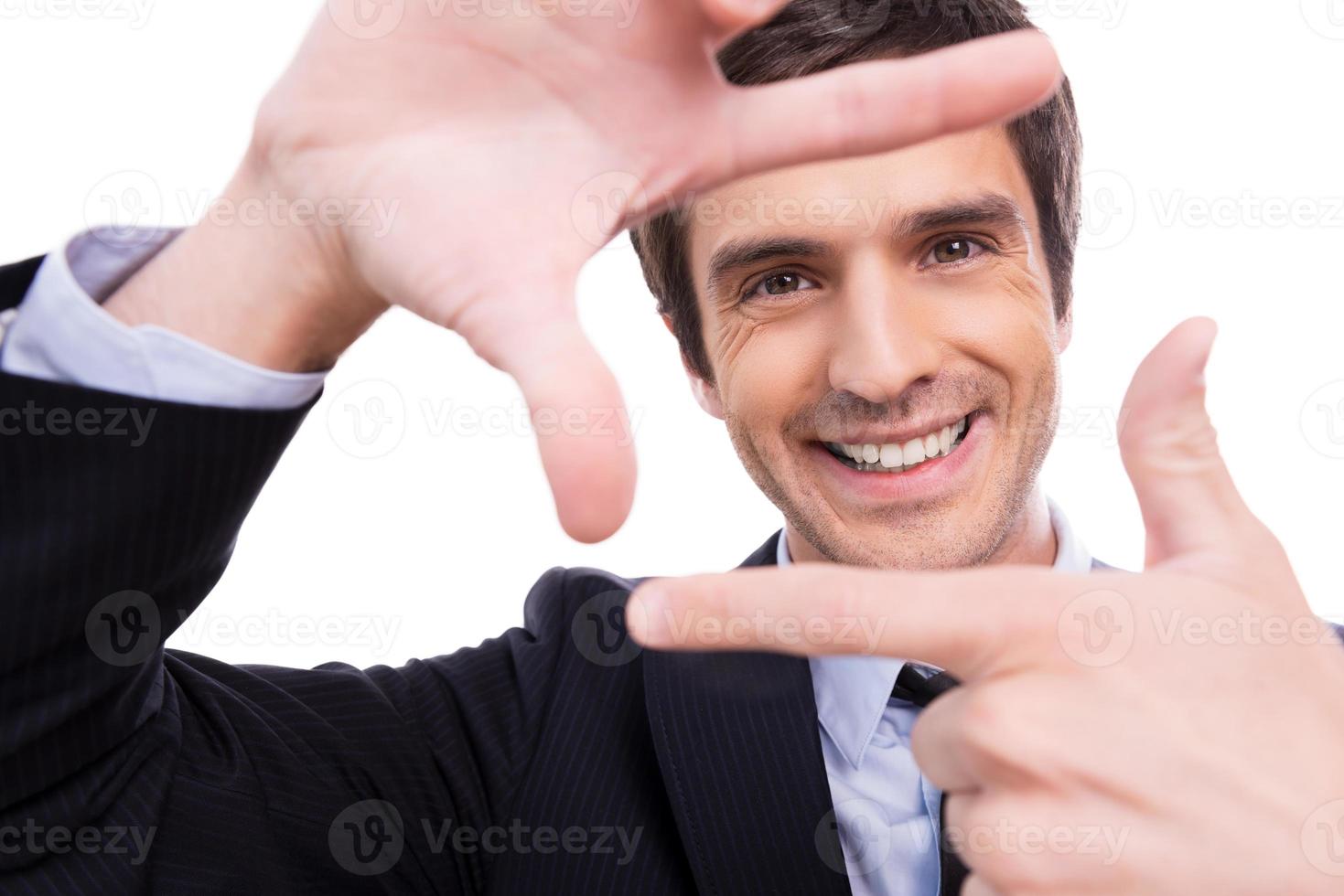 Focus on me Playful young man in formalwear gesturing finger frame and looking through it while standing isolated on white background photo