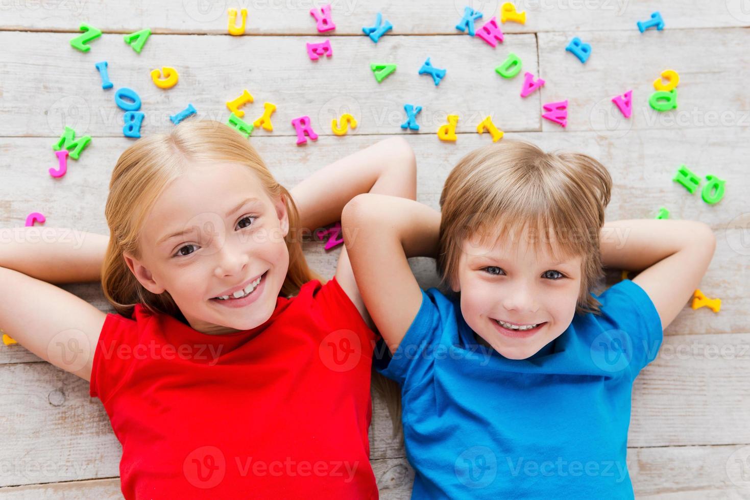 We love having fun Top view of two cute little children holding hands behind head and smiling while lying on the floor with plastic colorful letters laying around them photo