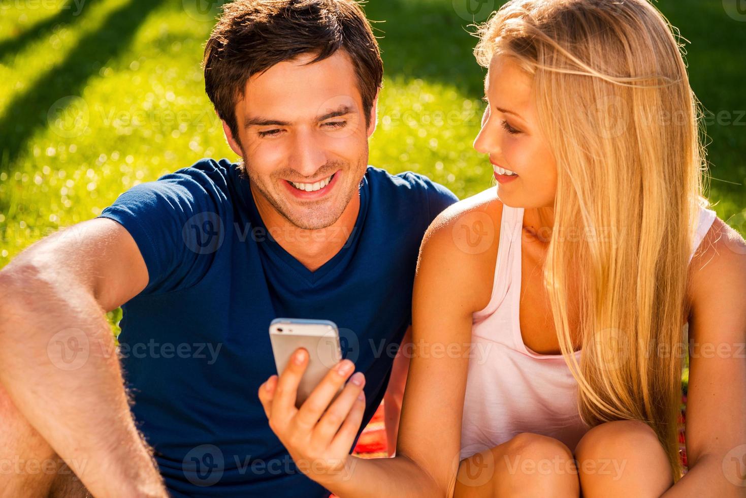 Look at this Happy young loving couple looking at mobile phone and smiling while sitting together on the grass in park photo
