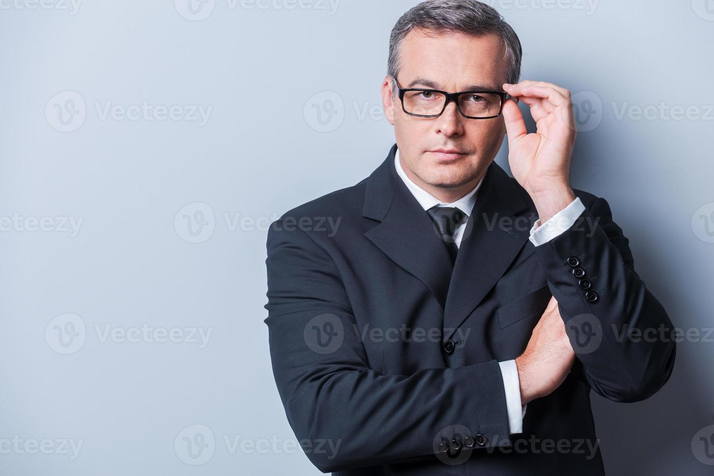 How may I help you Portrait of confident mature man in formalwear adjusting his eyeglasses and looking at camera  while standing against grey background photo