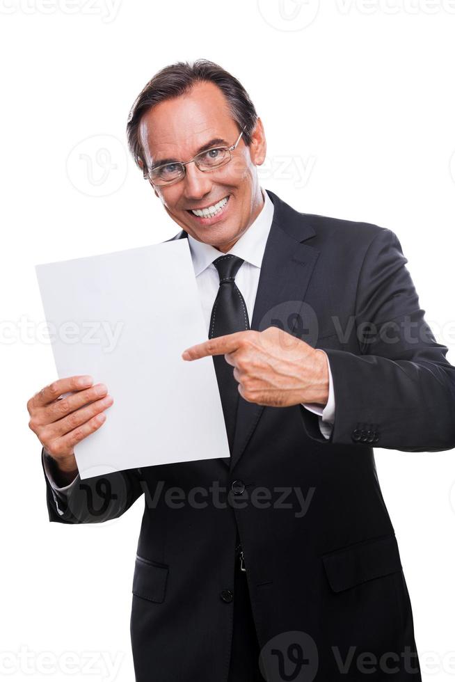 Your text here Happy mature man in formalwear holding blank paper and pointing it with smile while standing isolated on white background photo