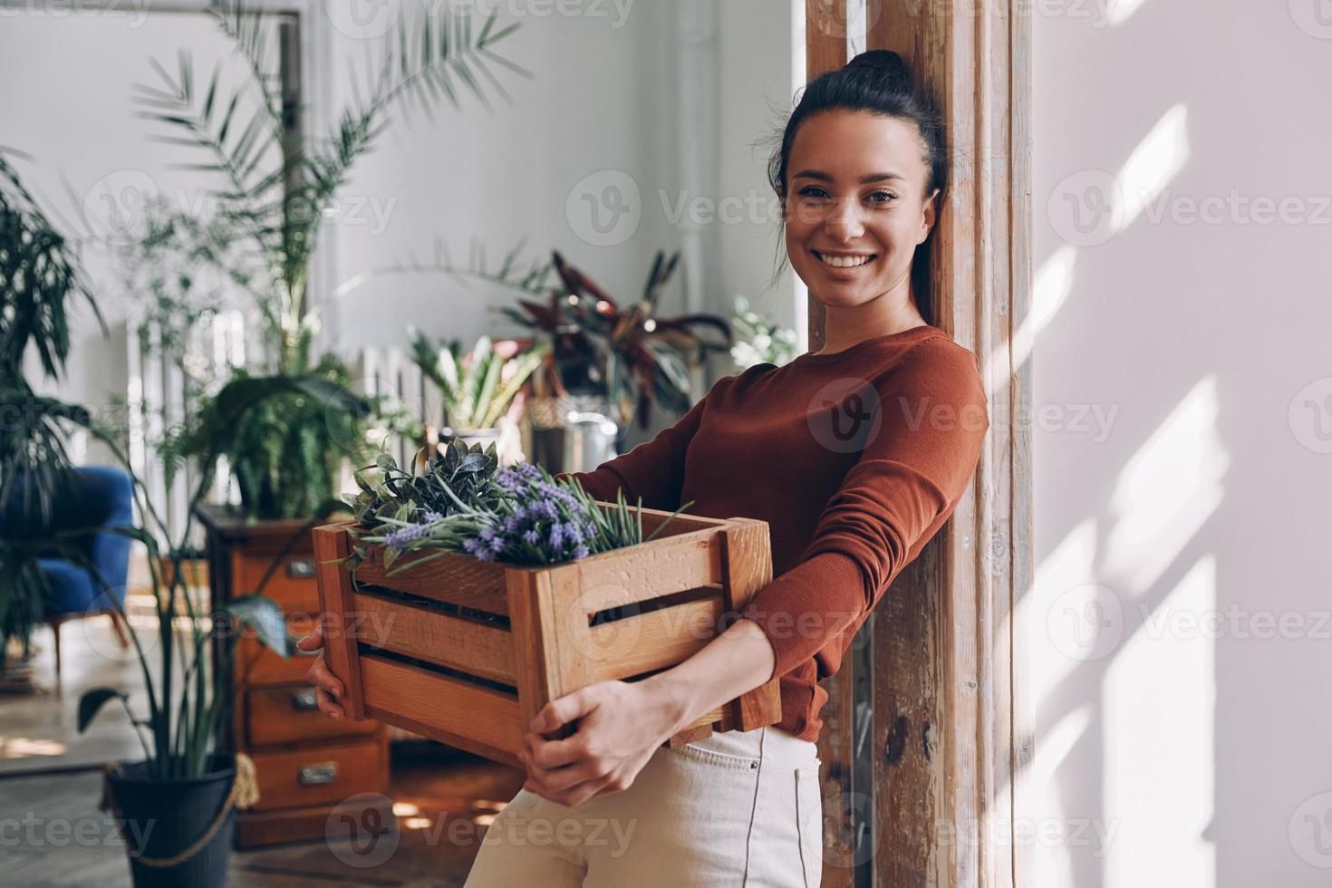 una joven alegre que lleva una caja de madera con plantas mientras se apoya en la puerta de casa foto