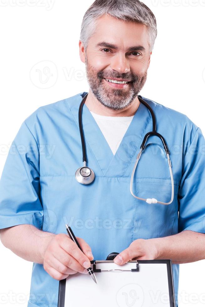 That is what you need Cheerful mature doctor looking at camera and smiling while pointing copy space on his clipboard and standing isolated on white photo