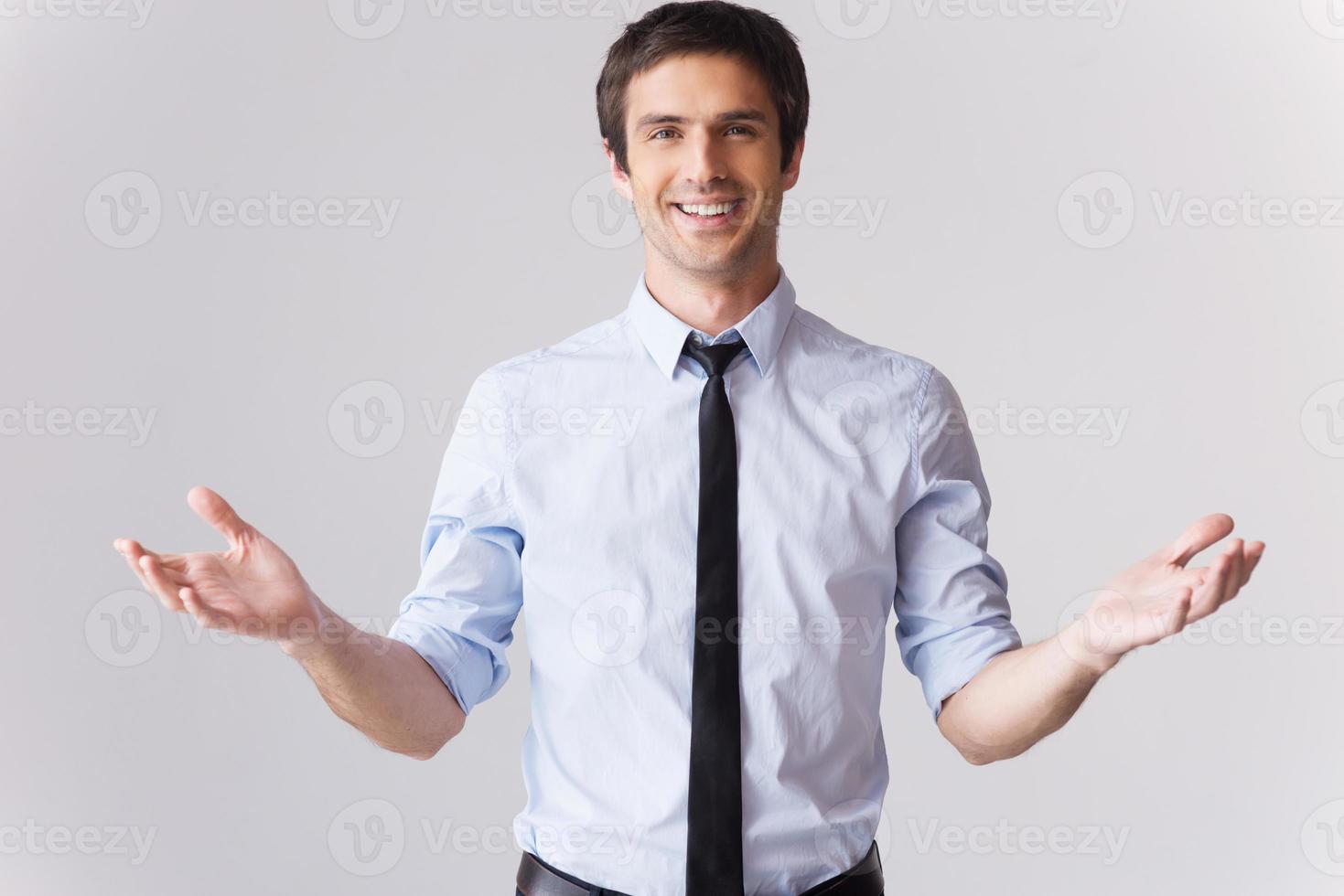 You are always welcome Handsome young man in shirt and tie gesturing and smiling while standing against grey background photo