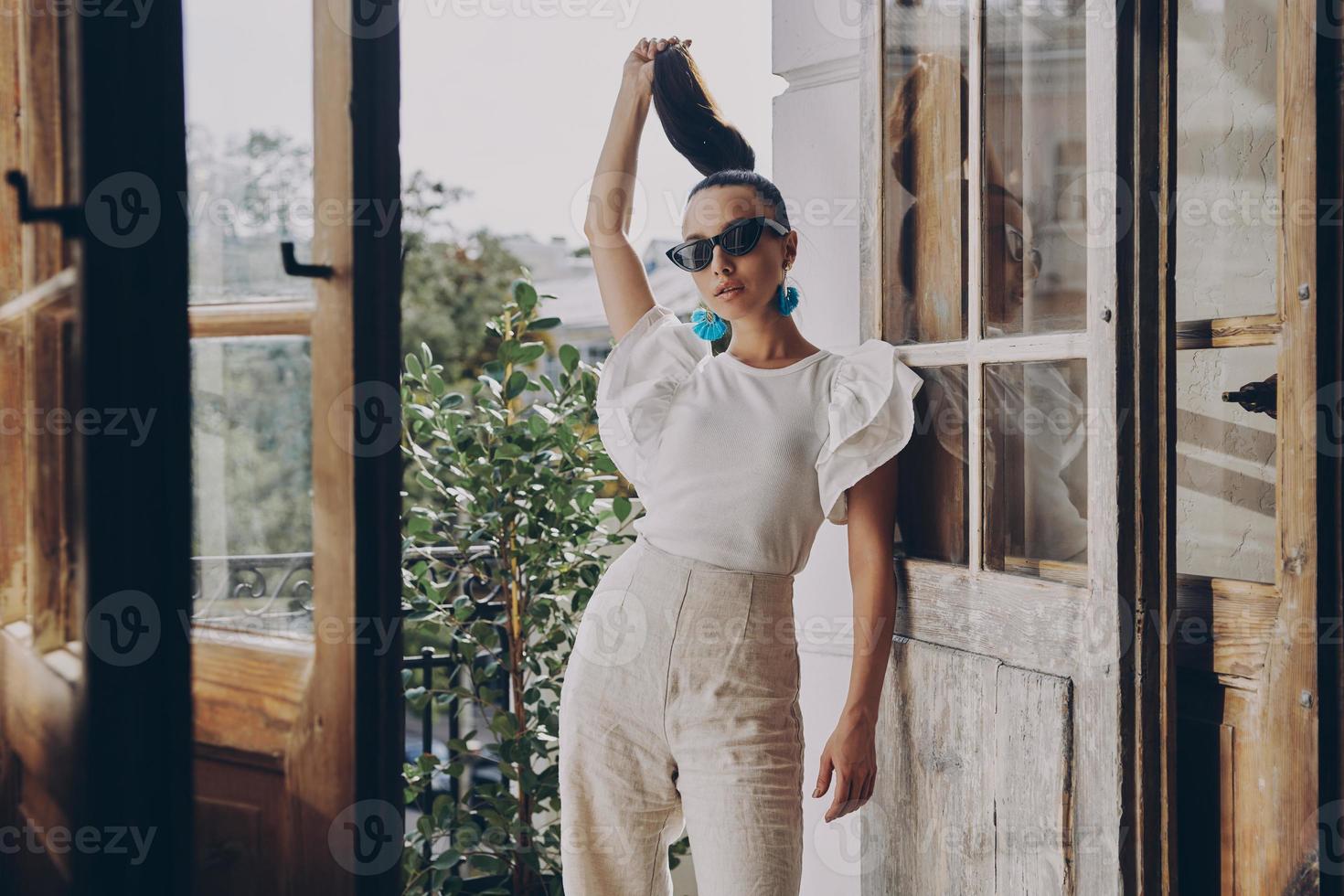 mujer joven de moda ajustando su cabello largo mientras está de pie en el balcón foto