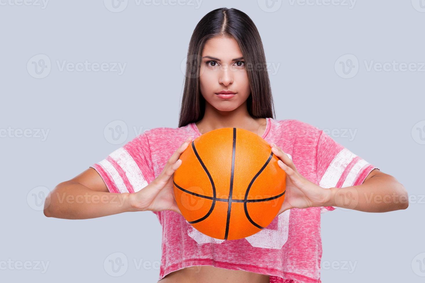 Are you ready to game Confident young female cheerleader holding basketball ball and looking at camera while standing against grey background photo