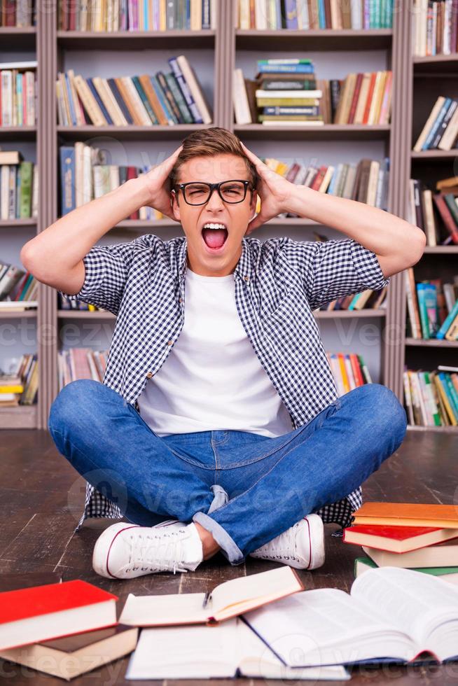 Tomorrow is my final exam Shocked young man touching his head with hands and shouting while sitting against bookshelf photo
