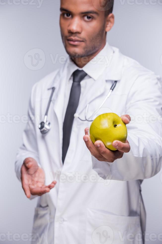 Join healthy lifestyle Confident African doctor stretching out green apple while standing against grey background photo