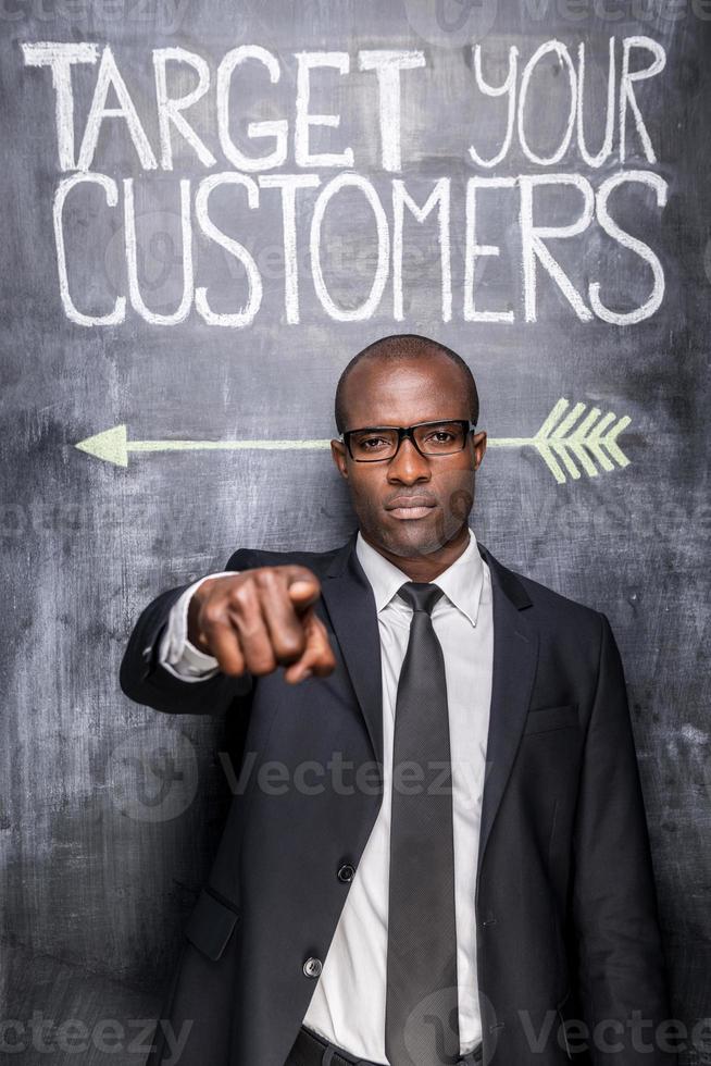 Target your customers Confident young African man in formalwear pointing you while standing against blackboard with sketch on it photo