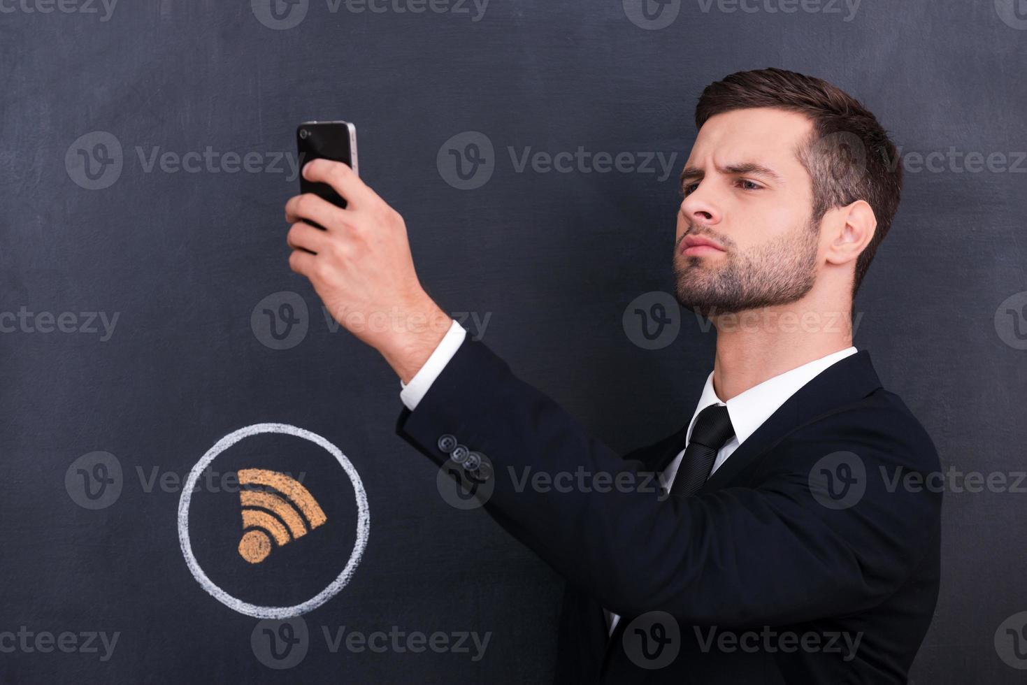 Where is Wi-Fi Picture of handsome young man holding telephone and looking for internet connection while standing against sharing  symbol chalk drawing on blackboard photo