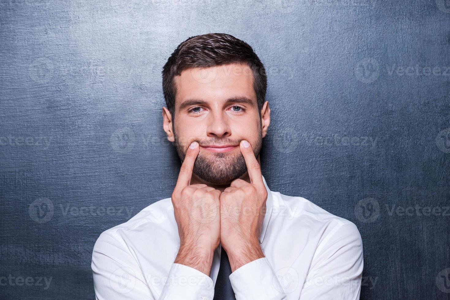 Put on your smile Handsome young man in formalwear holding fingers on his mouth and making fake smile while standing against blackboard photo